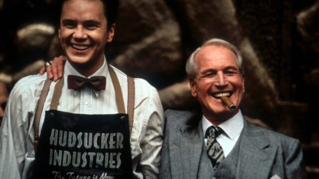 young man in mailroom apron beaming and standing next to older man in business suit with a cigar clenched between his teeth