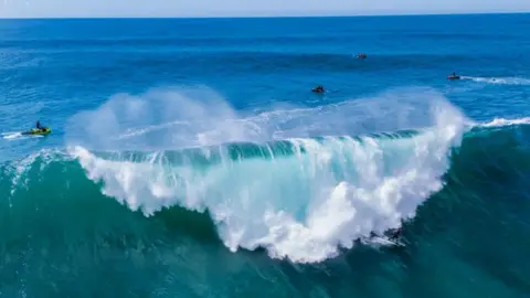 Getty Images A large wave in the Atlantic breaks. The water is a deep blue green colour. There is a surfer and people on jetskis. 