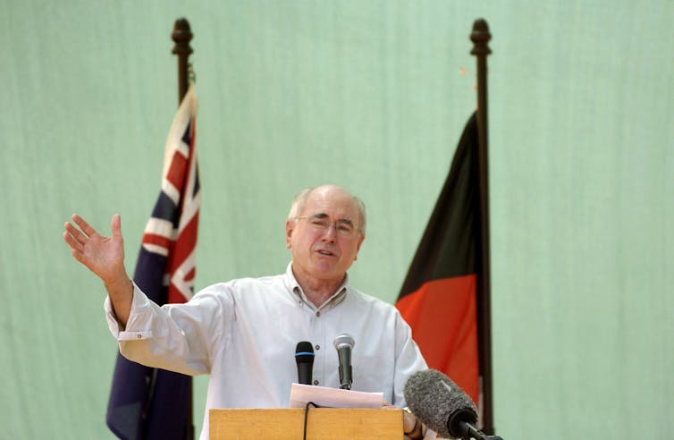 Former prime minister John Howard in a shirt with its sleeves rolled up at a podium in front of two flags