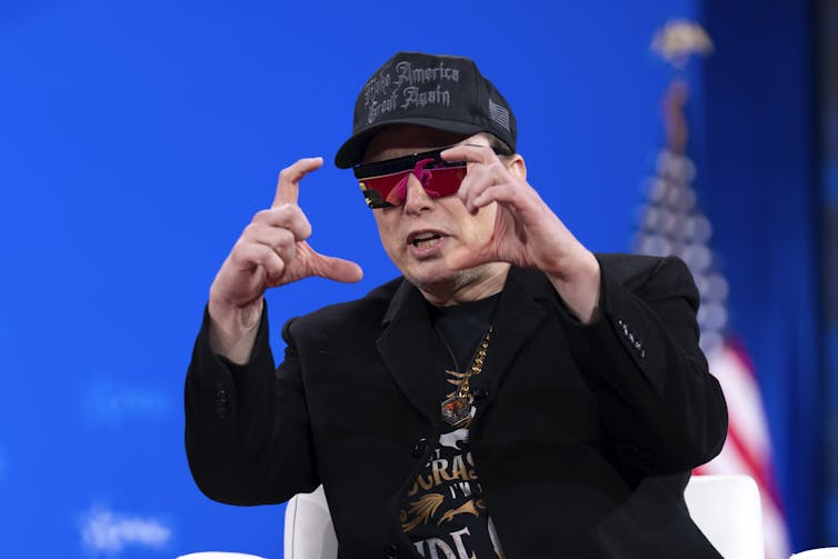 A man wearing reflective red sunglasses, a black suit and a black MAGA hat.