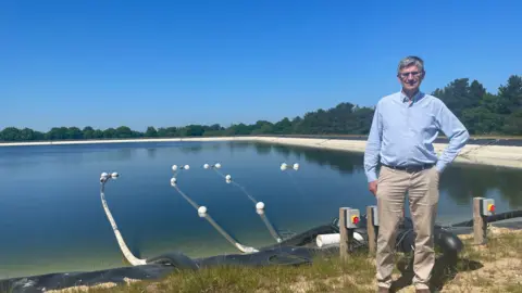 Malcolm Prior/BBC A man in a blue shirt and cream trousers stands next to a large reservoir with pipes leading out of it.