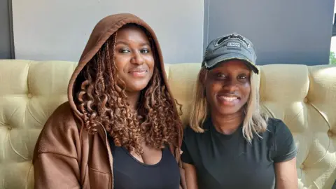 Hope Rhodes / BBC Two young black women sit in a booth at a bar smiling at the camera. Paris on the left wears a brown hoodie and Marie-Louise on the right wears a grey baseball cap. 