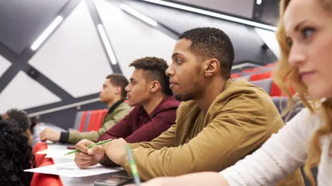 Getty Images A stock photo shows four students sat in a row of a lecture hall, as taken from the side. Three are men and there is a young woman in the foreground. They have papers in front of them and pens in their hands, and are looking intently to the left, towards the lecturer at the front.