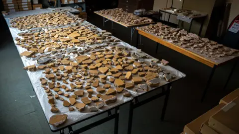 Tony Jolliffe/BBC News Hundreds of fragments of plaster laid out on several tables. Mainly yellow pieces making up three square panels with darker columns in between. 