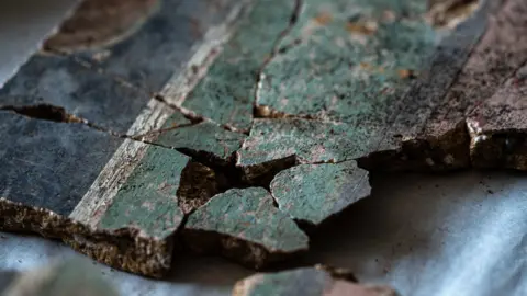 Tony Jolliffe/BBC News A close up image of at least 10 fragments of the plaster that have been pieced together. On the right hand side of the image is the pink dado section, then a thin black dividing line and a thicker pale green band. Towards the left of the image is a thin white dividing line and then a thicker black band. 