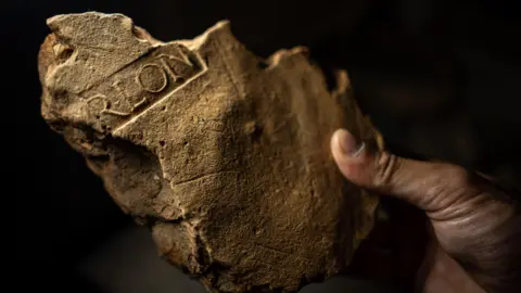 Tony Jolliffe/BBC News A close up of a broken piece of light brown stonework. Clearly visible are the remnants of some of the letters that have been imprinted in the plaster as it dried - R-L-O-N. It stands for Procurator of Londinium and shows that the building it was part of was a civil building in the growing city. 