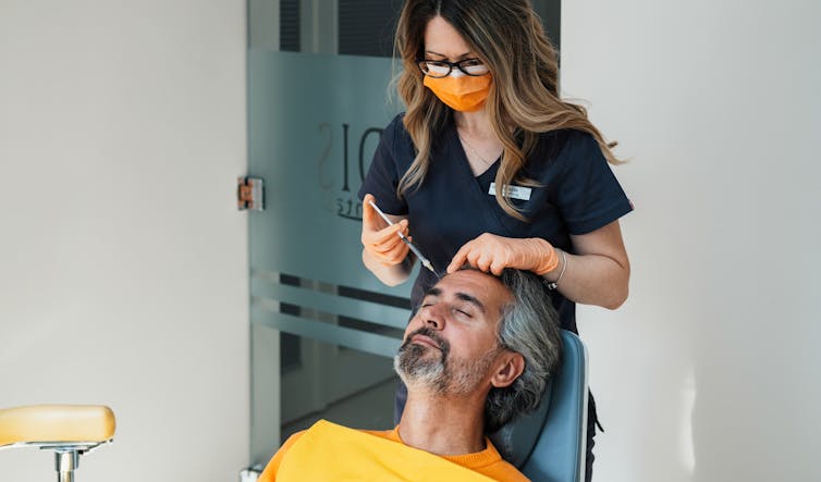 Masked woman injects forehead of a man lying in a clinic.