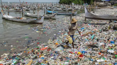Getty Images A person holding two plastic vessels walks across a shoreline full of rubbish, mainly plastics of various colours, with wooden fishing boats in the background.