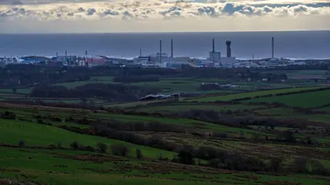 PA Media A general view of the Sellafield site seen from a distance, many chimneys are high over the site. There is the sea in the background and the clouds sit on the horizon. 