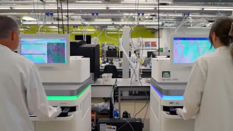 BBC News Two scientists facing away from the frame at two high tech white sequencing machines tha tlook like fridges with a computer screen on top