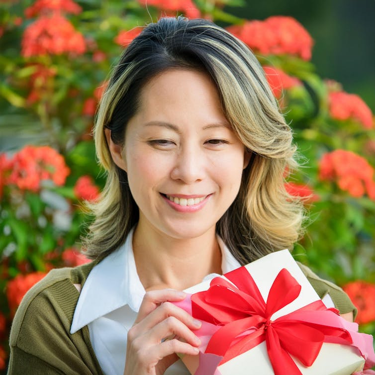 An Asian woman in a floral garden holding a misshapen present with a red bow.