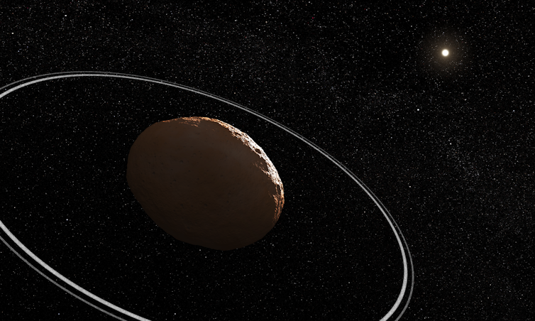 A rocky asteroid is in the foreground and a bright fuzzy dot representing the Sun is in the background. The asteroid has two narrow rings around it. The background is black and full of stars.