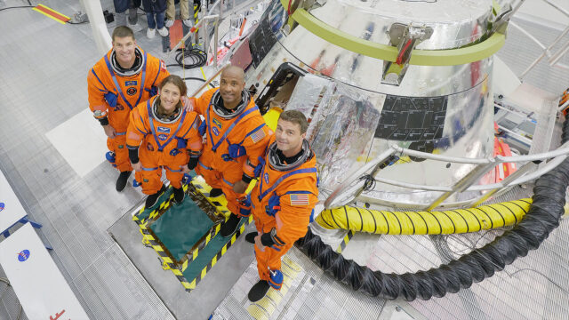 Three men and one woman, all in orange pressure suits, stand in front of a silver-coated space capsule in an overhead view