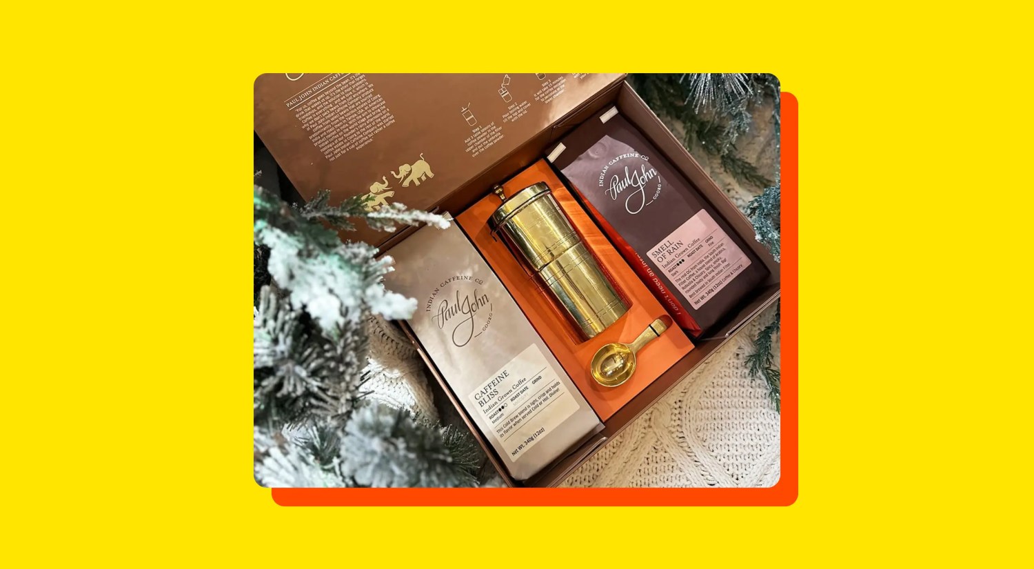 a photo of a box containing two bags of coffee and a brass coffee grinder and measuring spoon, bordered by a snow-dusted pine tree, on a yellow background
