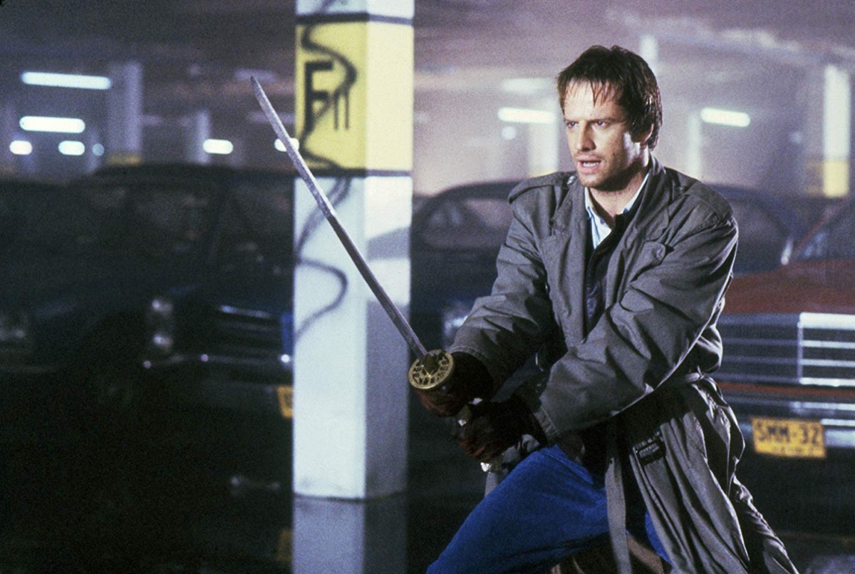 man in a trench coat wielding a vintage sword in a parking garage.