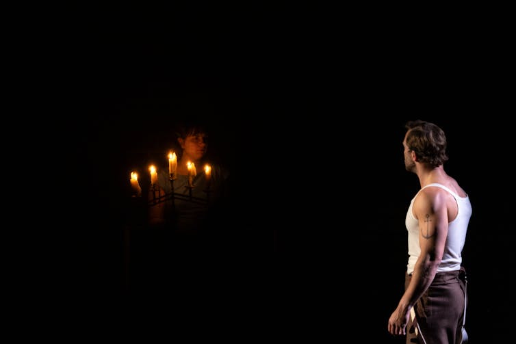 Production image: Tom looks at someone, half hidden, holding candles.