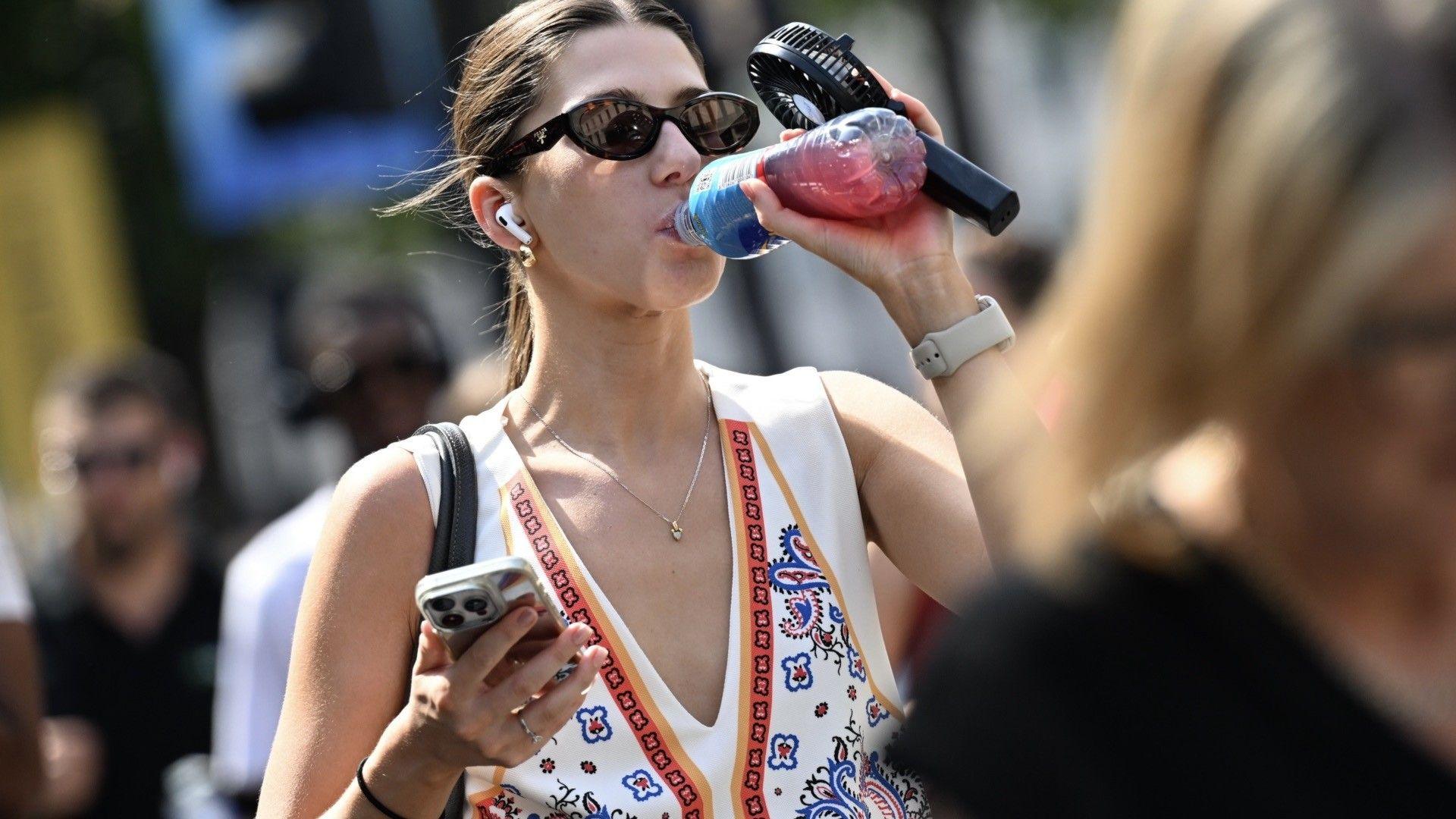 A woman drinks from a bottle, holding a portable fan, with a blurred picture of people in the background