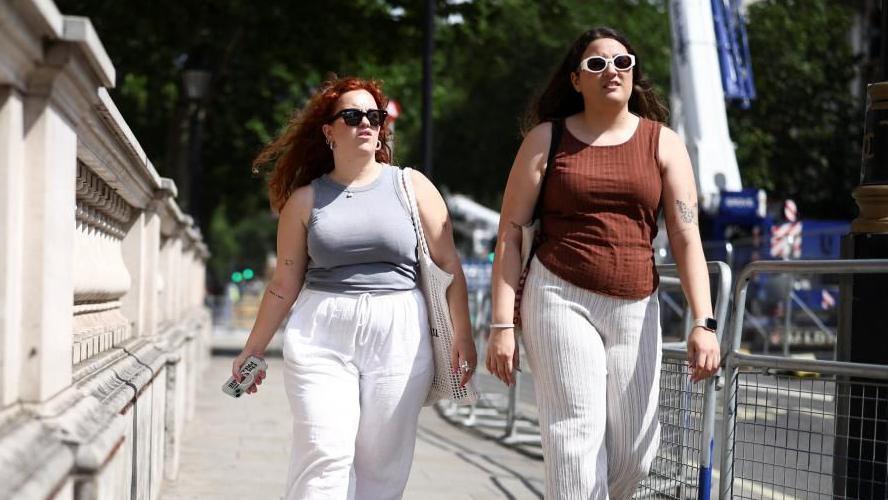 Two women walking through a street, with metal barriers to the right of them, wearing white trousers and vest tops - they both have sunglasses on