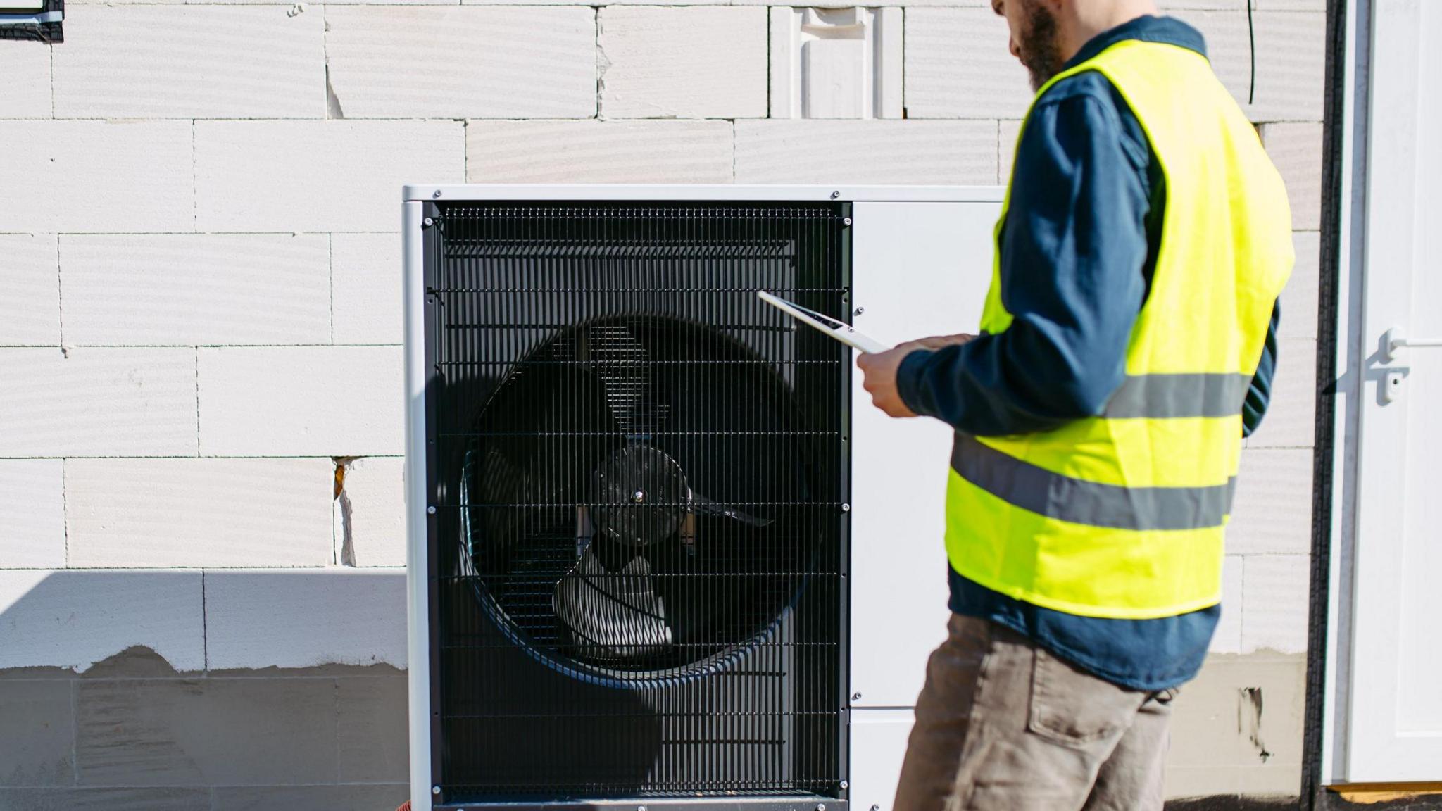 A black heat pump against a white breezeblock wall, a man stands to the right in brown jeans, blue shirt and a hi vis jacket holding a clipboard. He has a brown beard and the top of his head is cropped in the image