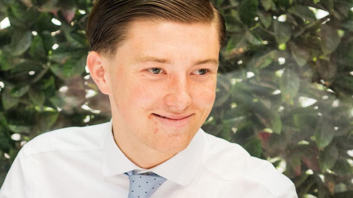 A young man wearing a white shirt and a blue tie. He is smiling. Henry has light brown hair. He is in front of a bush covered in green leaves.