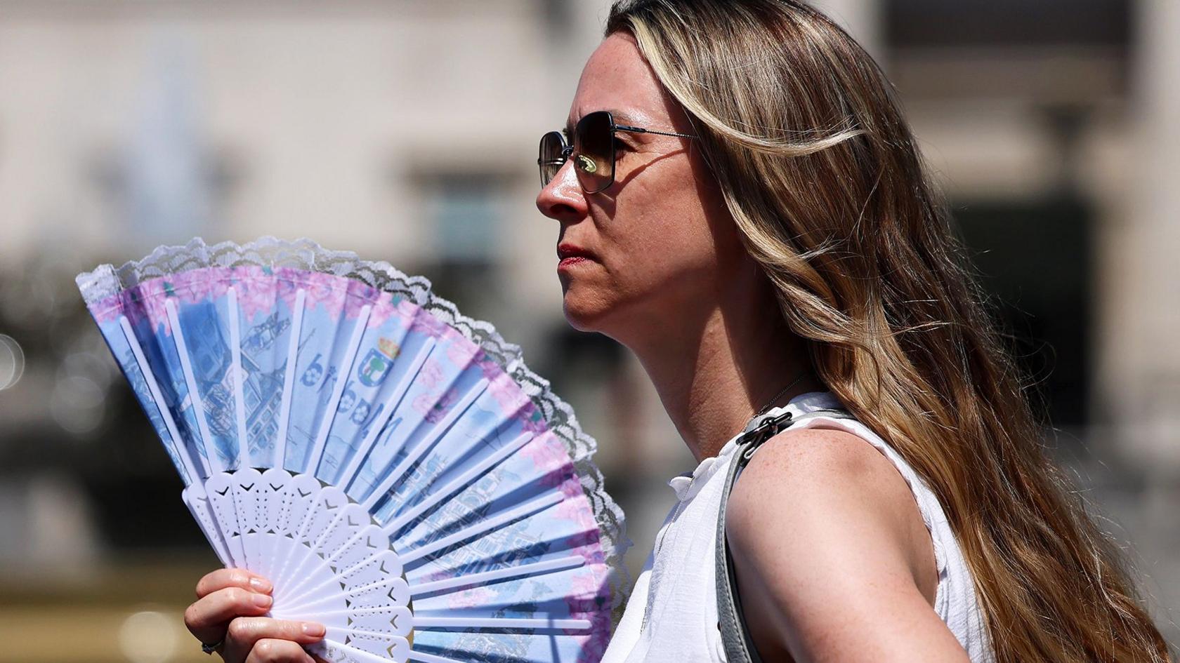 A woman in sunglasses wearing a white top holds a blue and pink fan in London on 24 June 2026