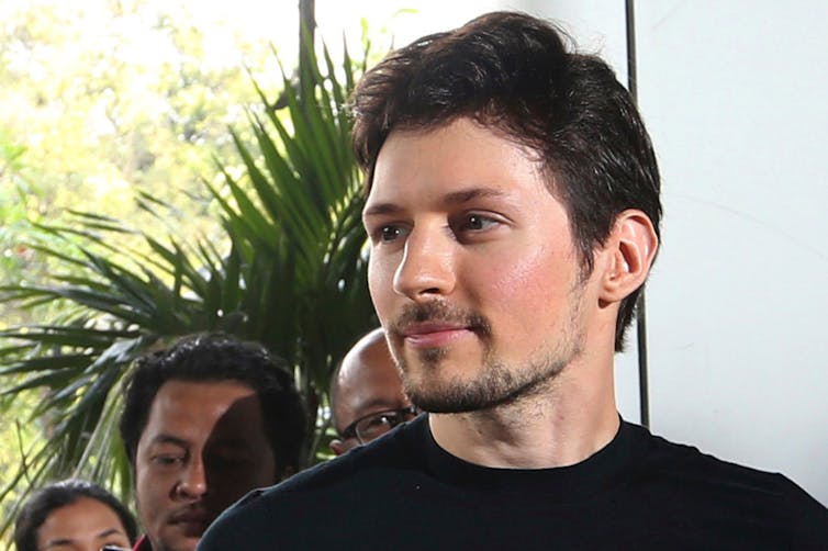 A white man with short dark hair, wearing a black shirt, standing in front of a group of people.