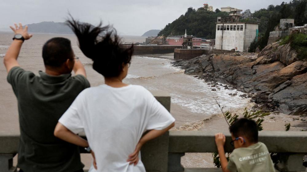 A man, woman and child stand behind a cemented barrier with backs to the camera, looking at muddy waters in a coastal region. There is a rocky shore and a few buildings to the right. The woman's hair is tossed into the air and the child is holding on to the barrier.
