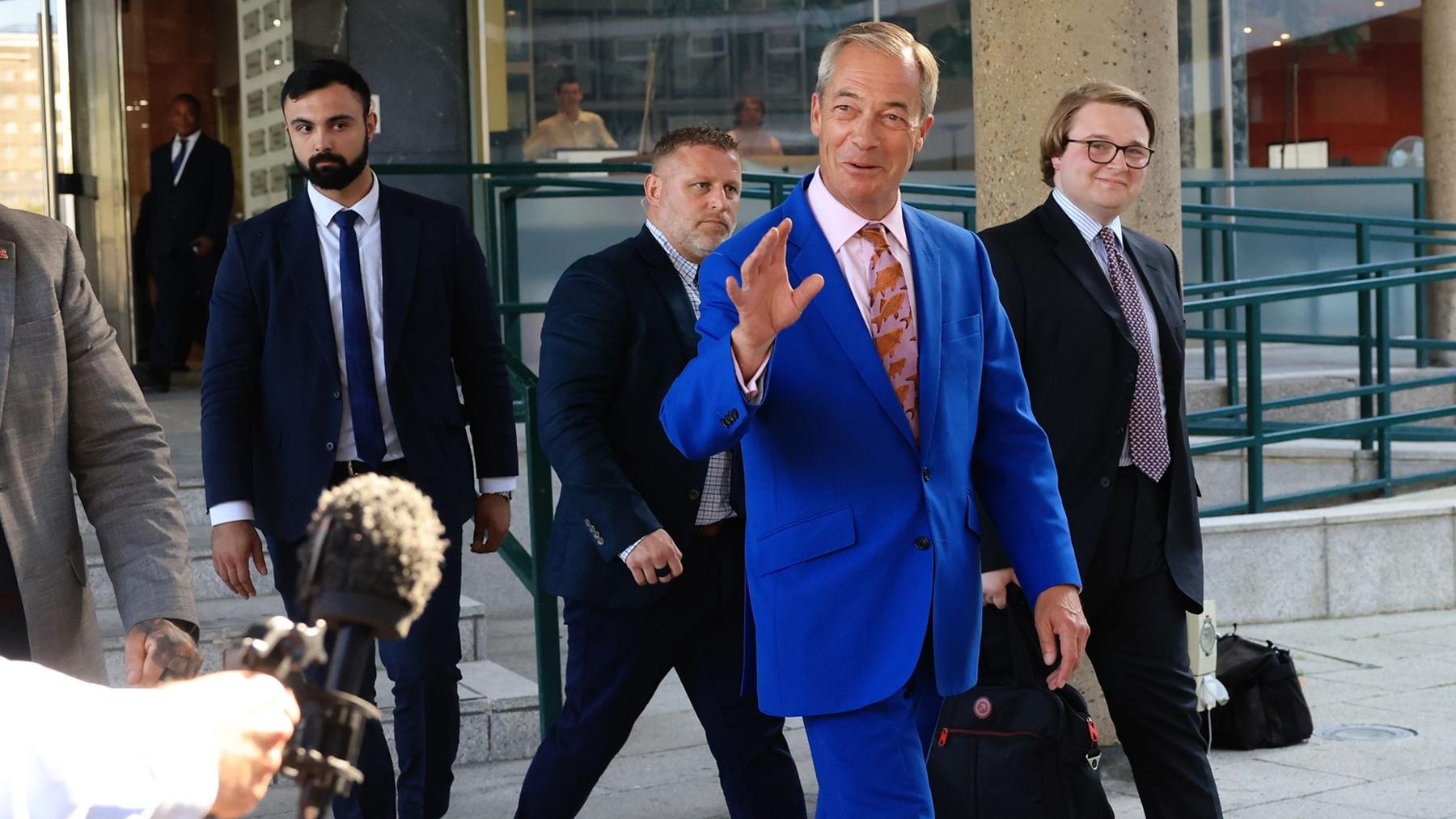 Nigel Farage walking outside, wearing a blue suit with a pink tie, decorated with a fish pattern. There are two men behind him, both wearing dark suits.