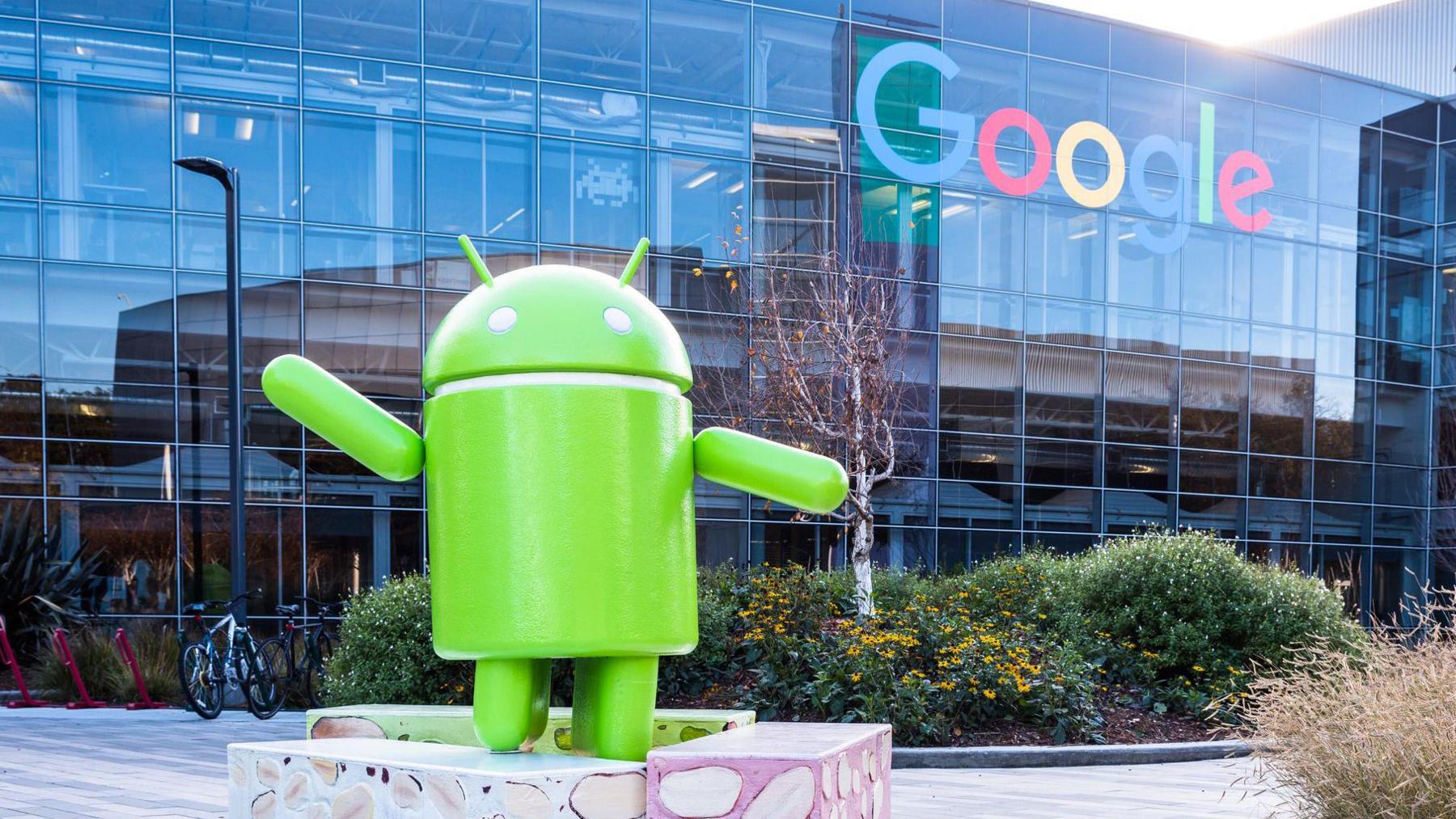 The green Android logo as a sculpture placed outside a glass Google office building, which has the Google logo on it.