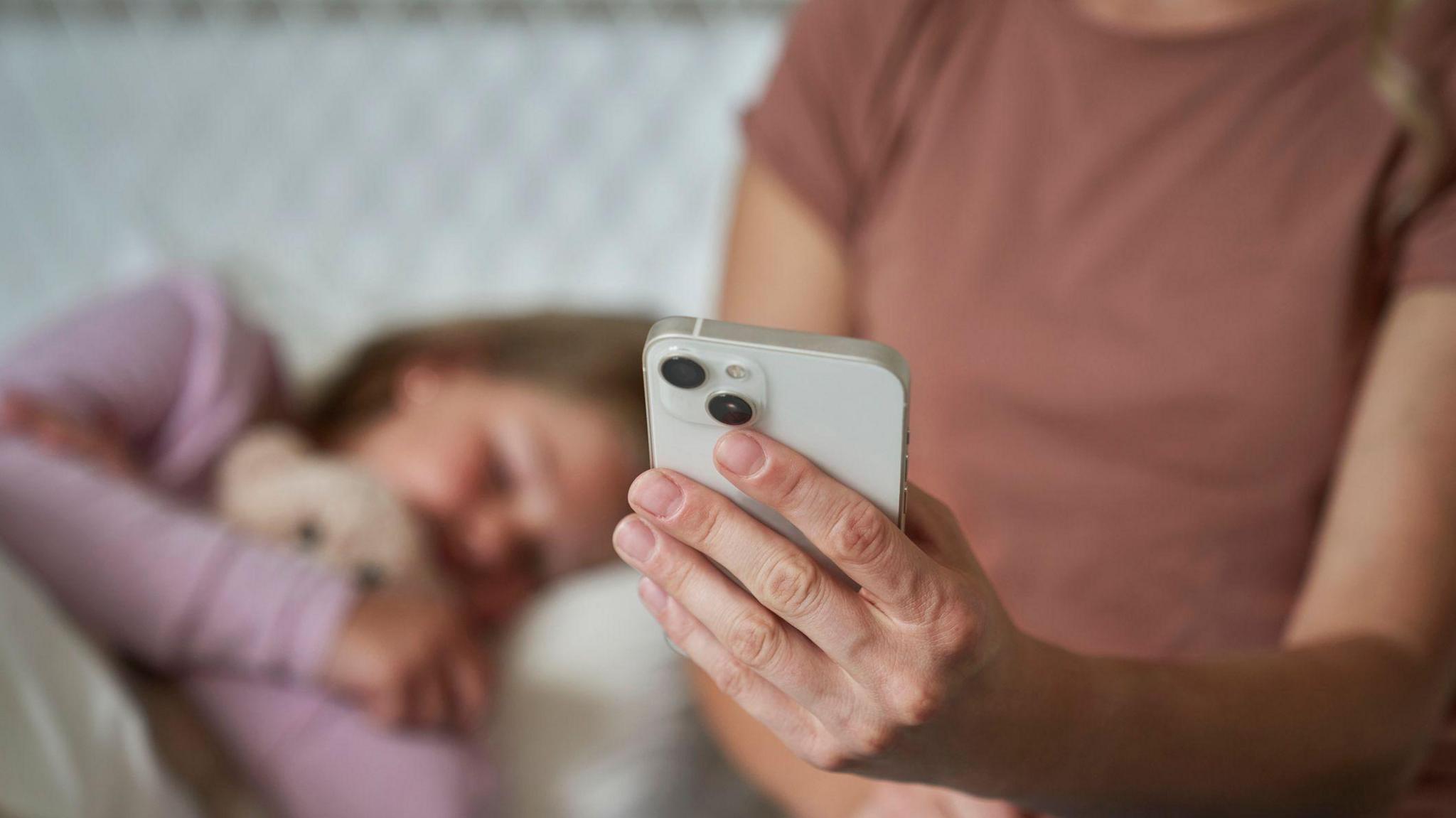 A mum, whose face cannot be seen, holds a smartphone out in front of her while her young daughter lies in bed beside her. The child's face is blurred.