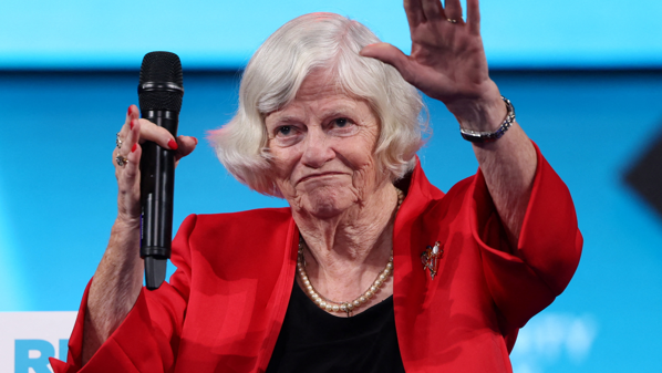 Ann Widdecombe waves on stage - she has a microphone in one hand and is wearing a red jacket, black top and pearls