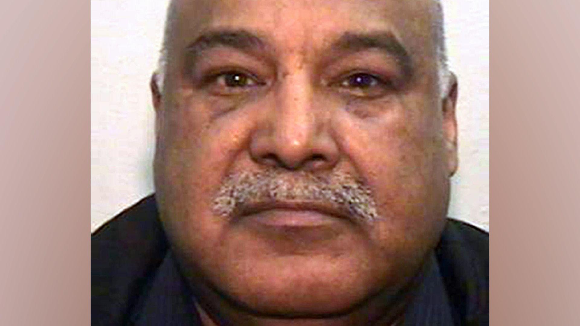 A mugshot of an Asian man in his 60s, with a grey moustache and bald head. He is looking into the camera.