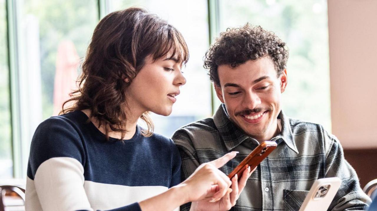 A woman with dark shoulder-length hair points at a smartphone held by a smiling man with curly hair and a moustache.