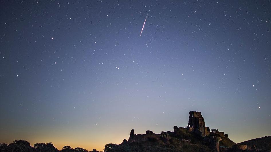 A bright Perseid meteor streaks across a star-filled night sky above the silhouette of Corfe Castle