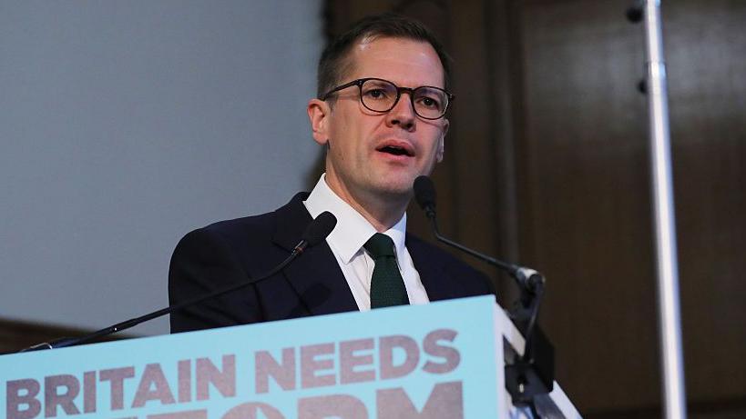Headshot of Jenrick who speaks into two small microphones on stands on a podium in front of him that has a blue sign partially cut out of the frame, but you can read it says'Britain needs reform'.