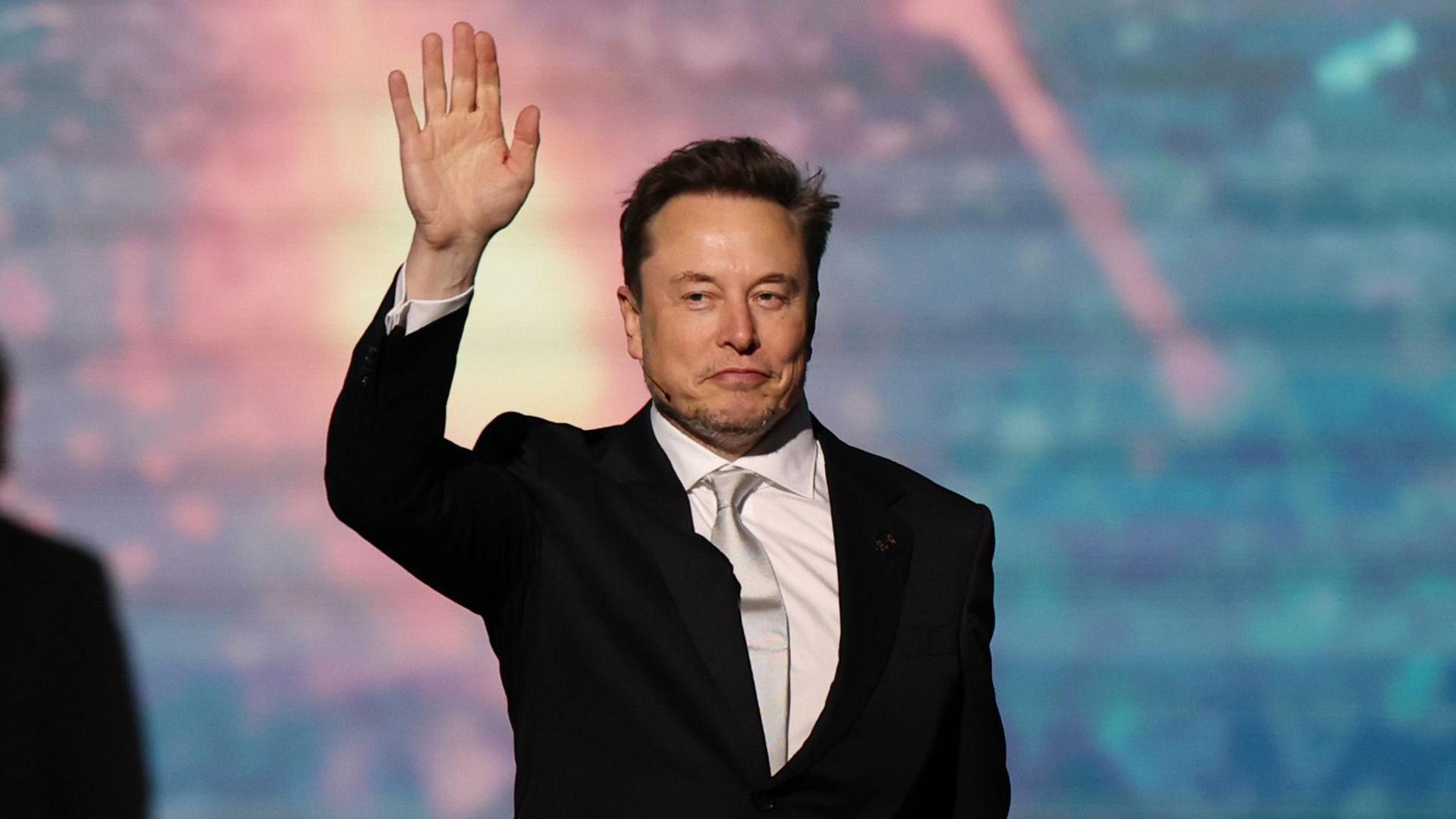 SpaceX chief executive Elon Musk walking onto a stage and waving, wearing a black suit, white collared shirt and shiny off-white neck tie.