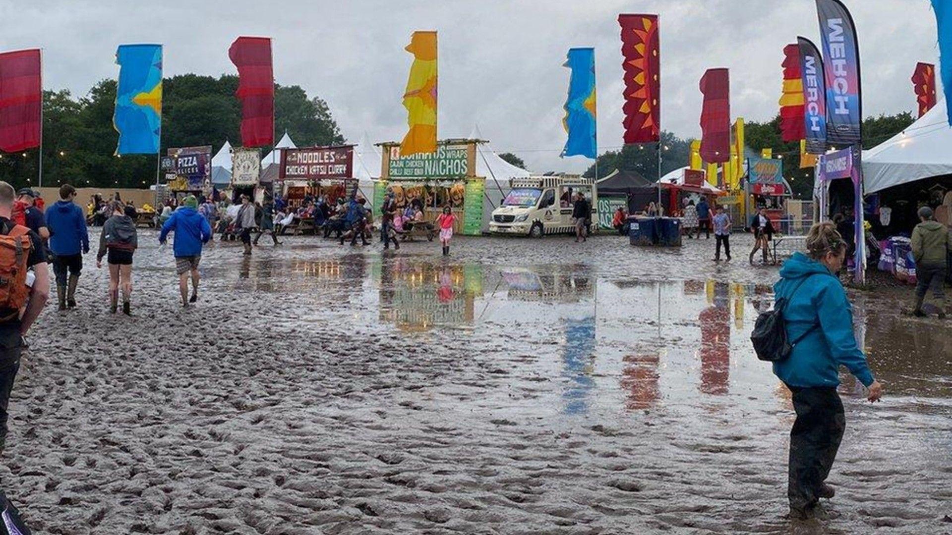 Muddy conditions at a festival site with people wearing raincoats and wellington boots among flags and marquees flapping in the wind