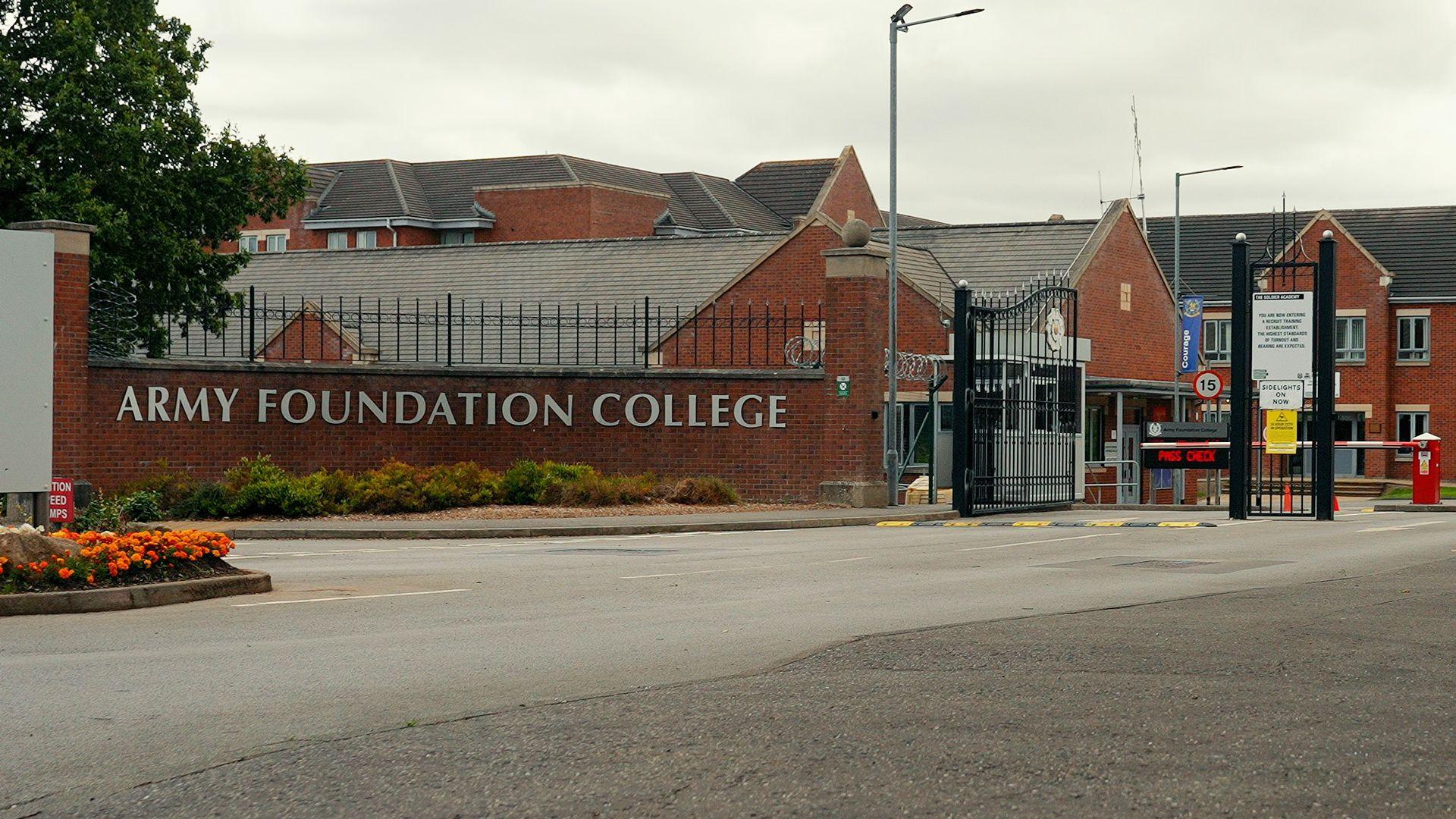 The words Army Foundation College are on a brick wall at the entrance to the campus. Large black gates are open with a red and white barrier slightly beyond. The college's buildings are behind.