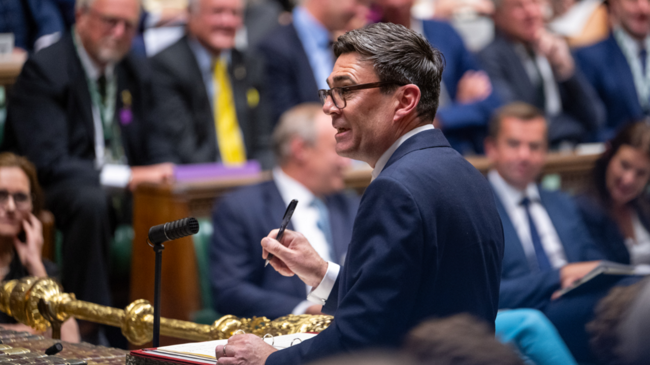 Andy Burnham speaking in the House of Commons. He is wearing a dark suit and holding a pen.