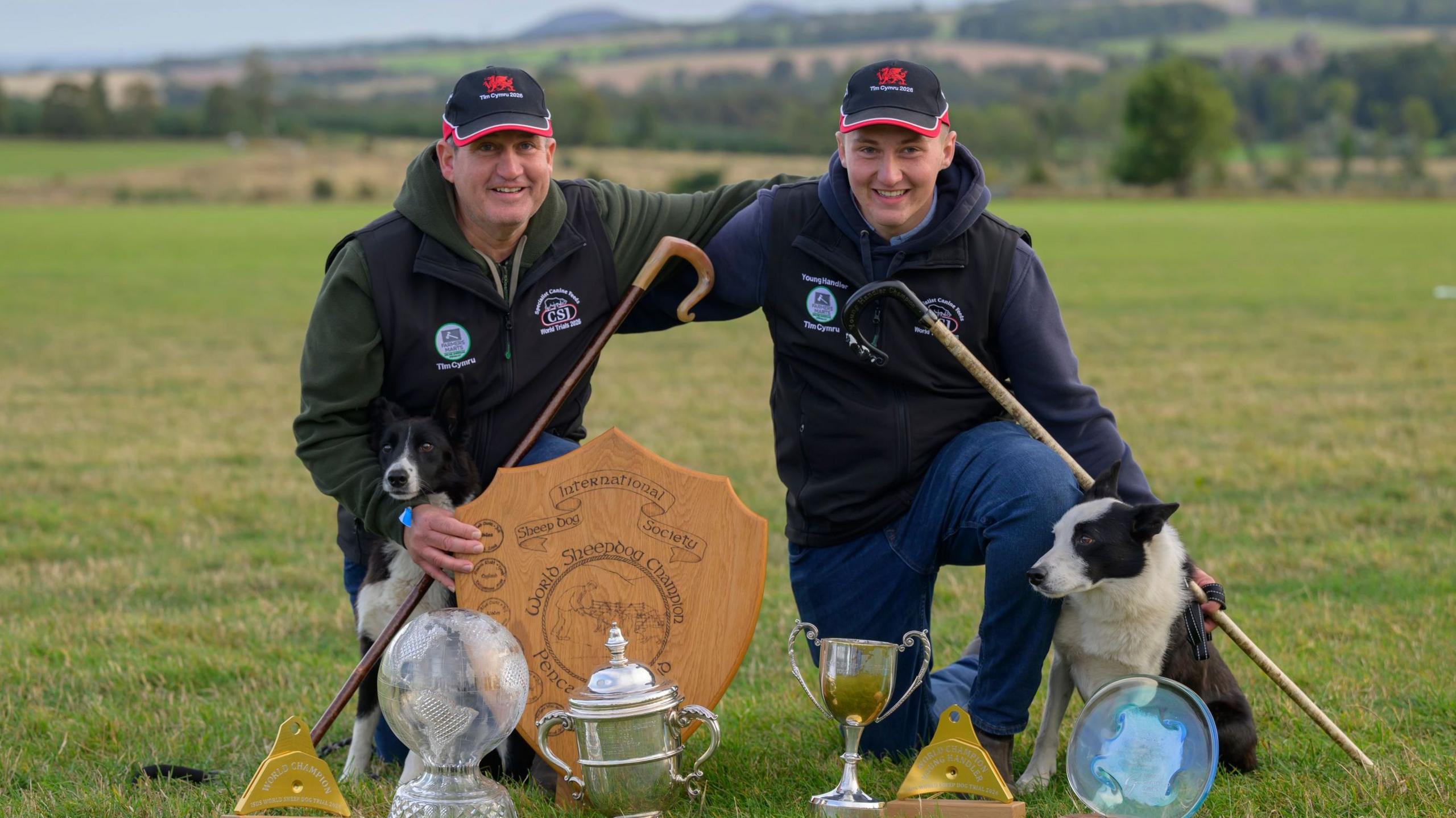 David a Jack Howells in a field on their farm, crouching down and smiling, with their arms around each other. They have several trophies in front of them, and their dogs are also in the photograph. They are both wearing black baseball caps with a red Welsh dragon.