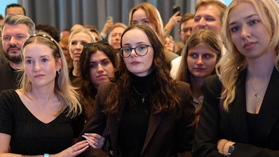 More than a dozen CDU supporters dressed in formal wear are seen with neutral expressions looking at screens. A woman in the front folds her arms, while another holds onto the arm of the woman next to her.
