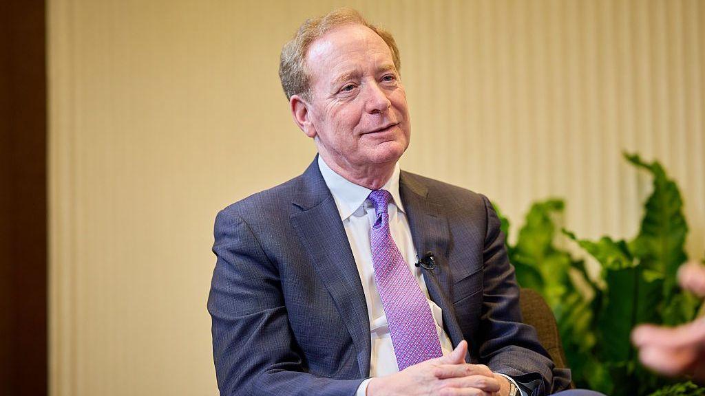Microsoft president Brad Smith sitting with his hands clasped in his lap wearing a blue suit, white shirt, and purple-patterned tie. Behind him in soft focus, there is a beige background and some green plants.