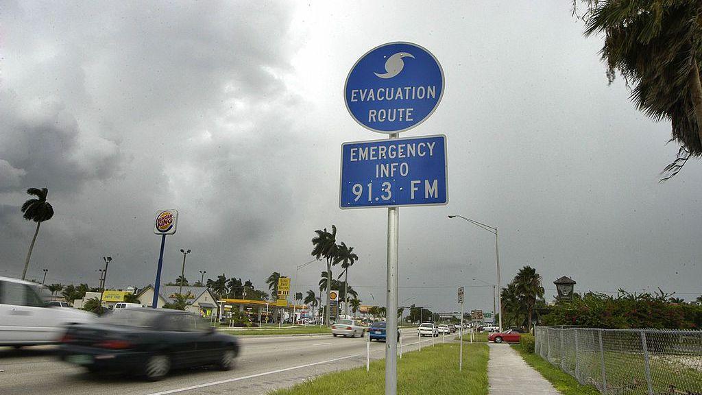 Photo shows a road with cars passing a blue sign that has a hurricane symbol and says'evacuation route''emergency info 91.3FM'