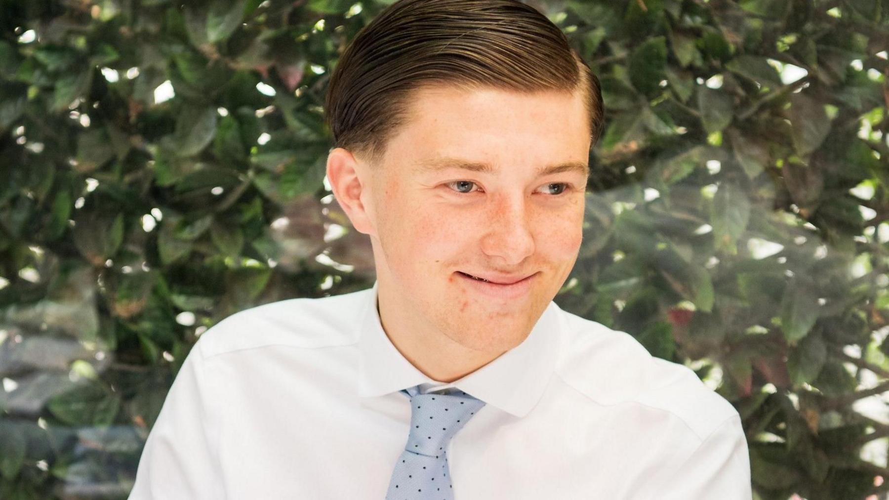 A young man smiling wearing a white shirt and a blue tie. He is sitting in front of a green bush. He has brown hair.