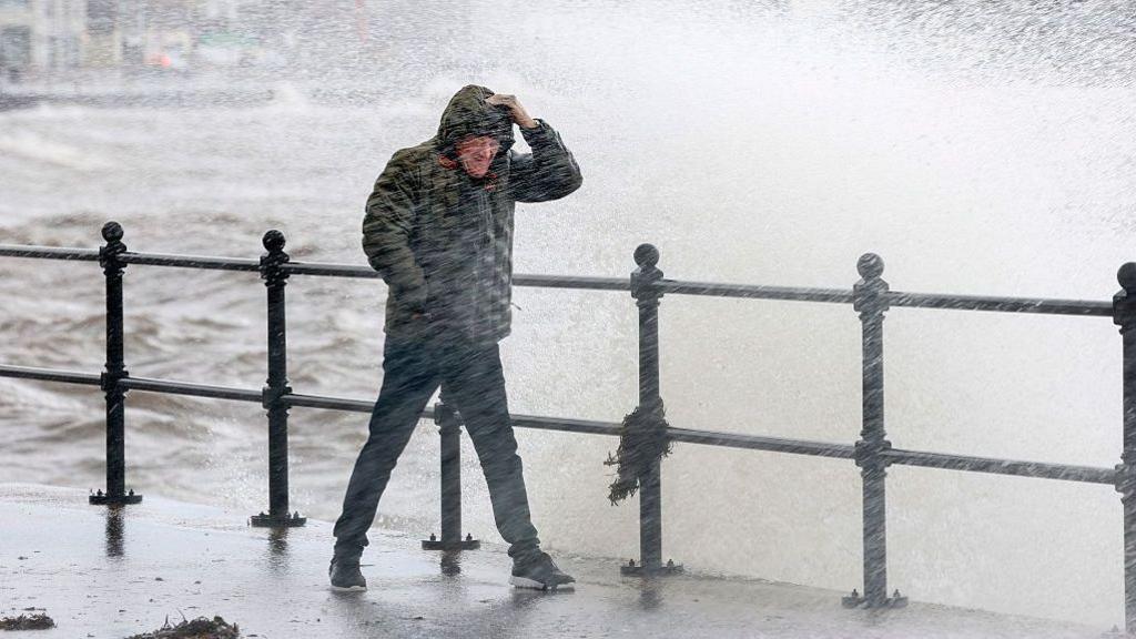 A man walking near the coast. He is wearing dark clothes and a coat with a hood up, and is being drenched by a wave which has come up and over the black railings.