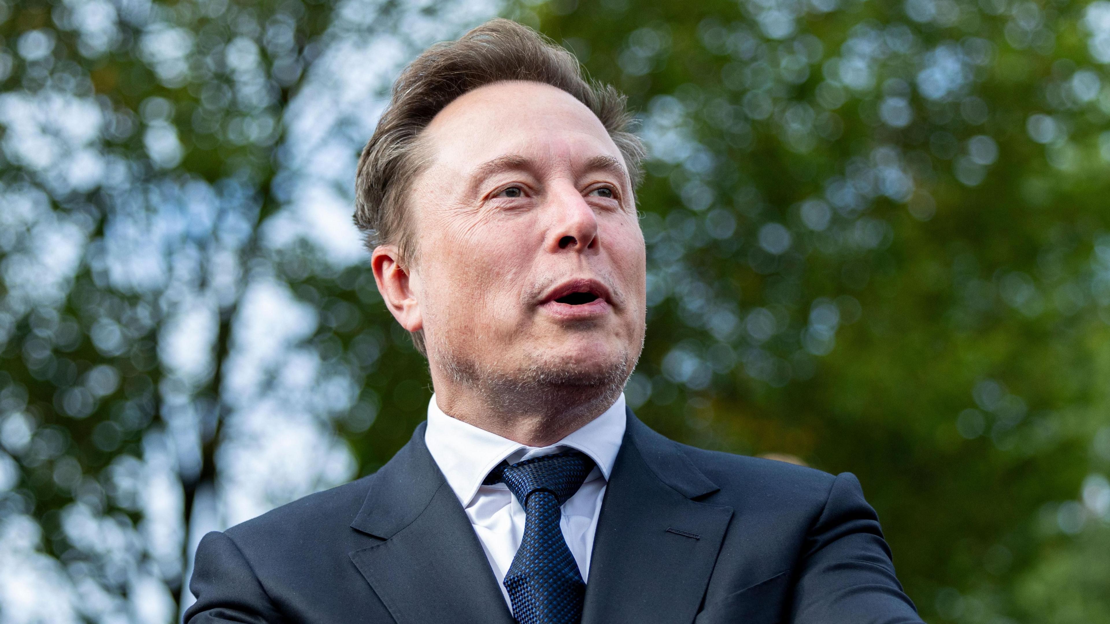 Elon Musk speaking and looking to the right with leafy green trees in the background.