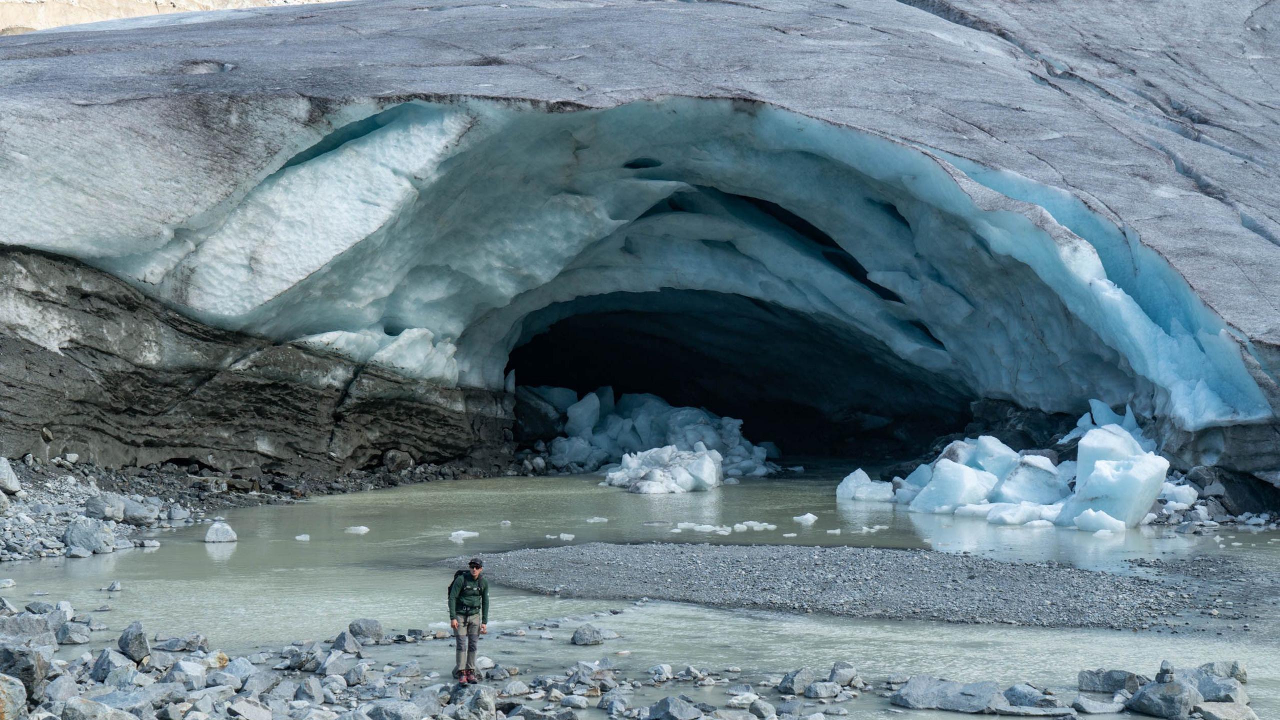 The front of a glacier, where a large channel - which looks a bit like a cave or an arch - has eaten away at the ice. A river of meltwater now meanders out of the front of the glacier. There are also chunks of ice on the ground having broken off the glacier. A man, dressed in mountaineering clothing, stands in front of the glacier for scale, which shows how big the channel or cave is.