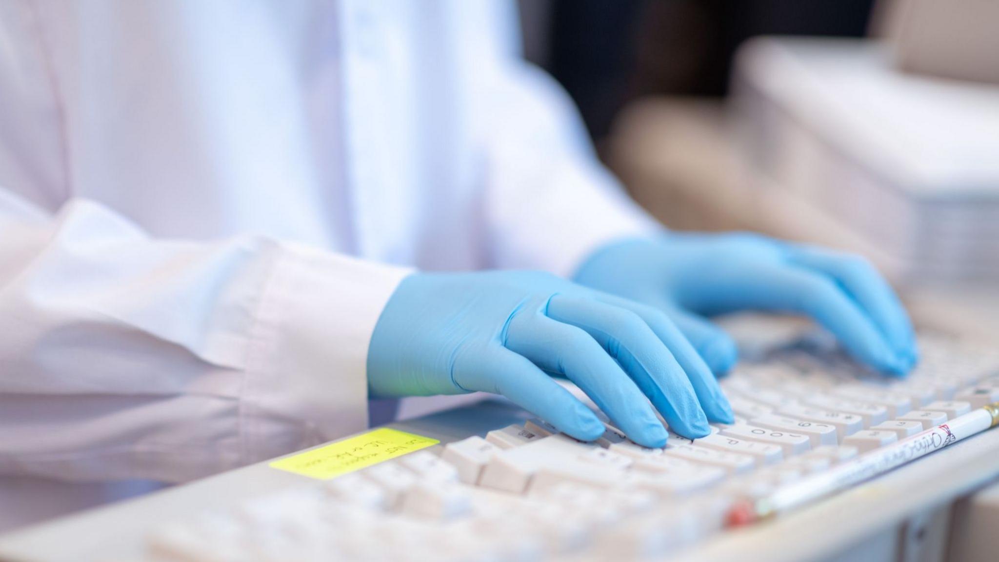 A close-up stock image of a lab worker, wearing a white coat and blue gloves, typing on a keyboard.
