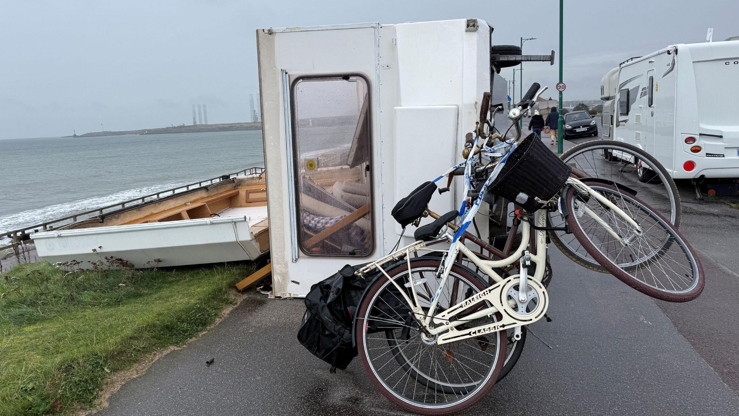 A caravan is blown in its side on a beachside road. It has two bikes attached to the back