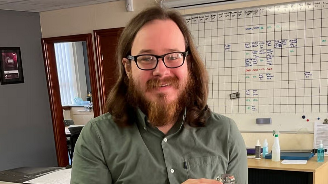 A photo of Joshua Kerry, a man with shoulder-length brown hair, a beard and glasses, from the shoulders up. He is smiling at the camera, wearing a khaki shirt, and behind him is a whiteboard.